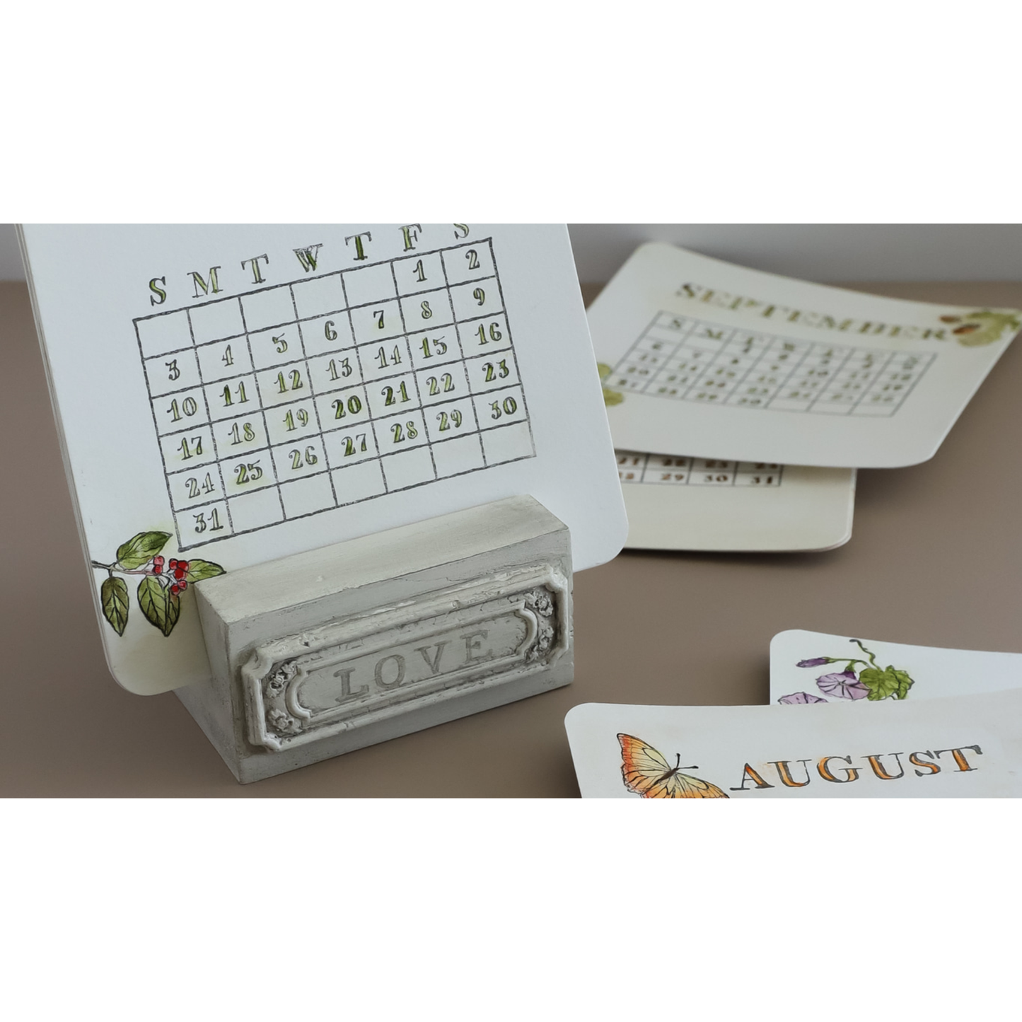 A small tabletop calendar with a white card showing the weekly schedule and a decorative gray base engraved with the word LOVE. Additional calendar pages and printed cards are arranged on a beige surface.