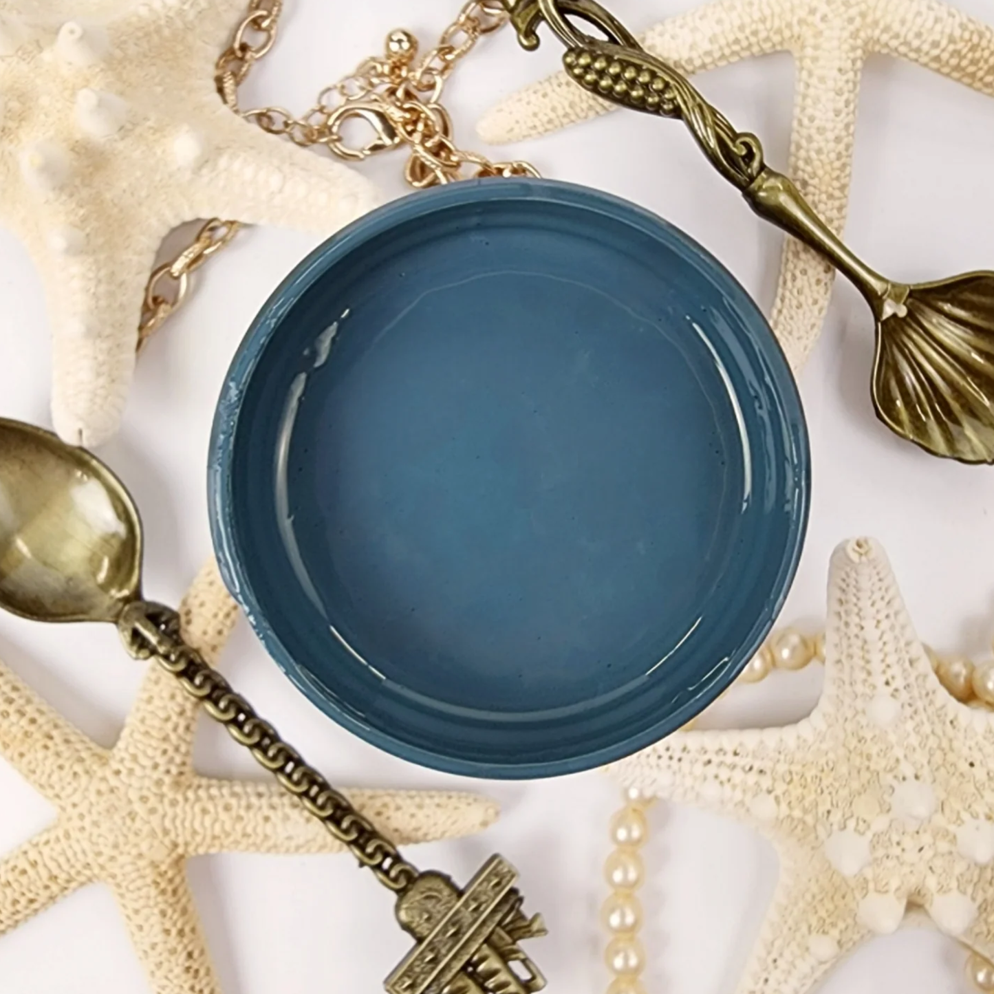 Image of open jar of deep blue/green colored paint with brass spoons, starfish and tarnished pearls on a white background.