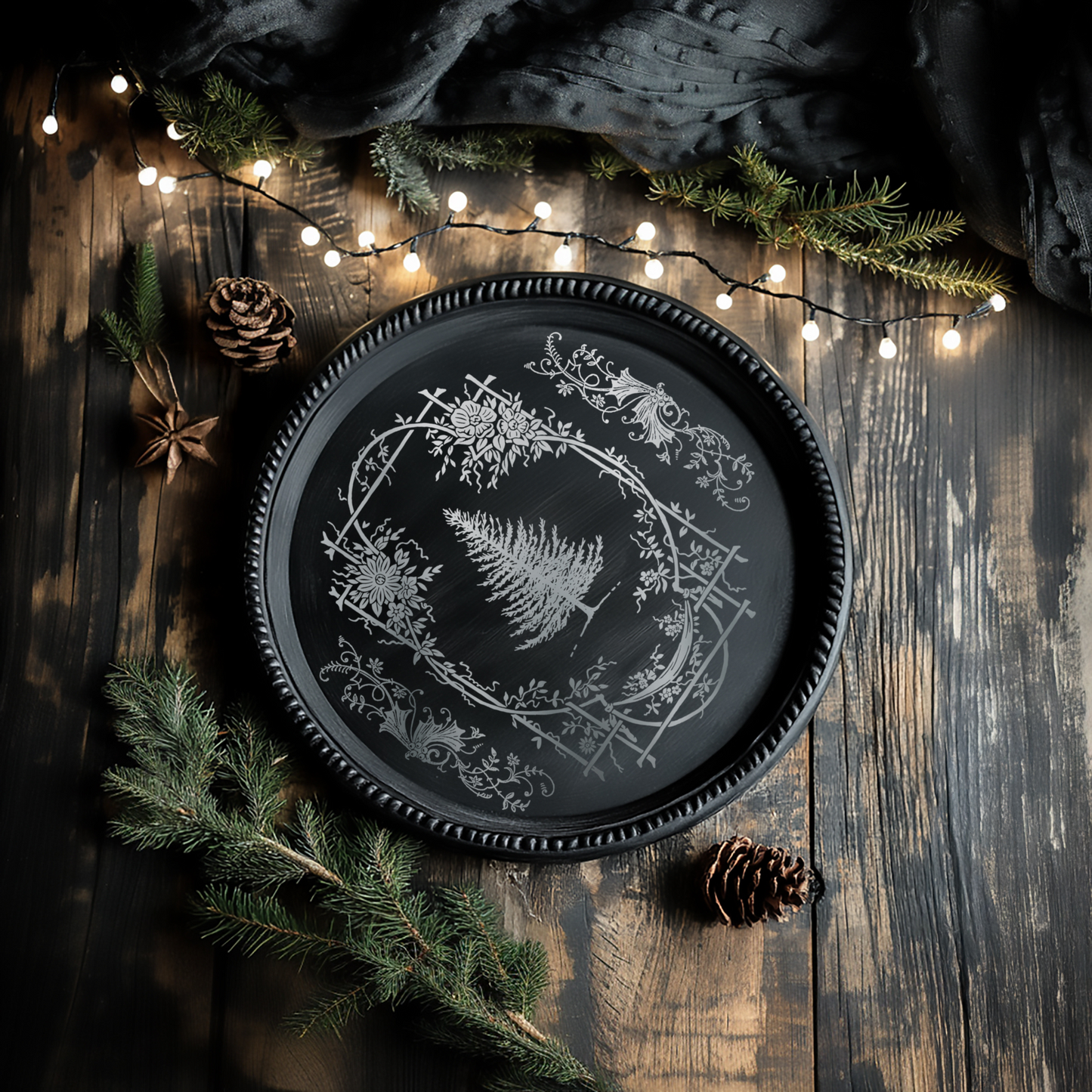 "Figgy & Flourish" IOD Gilded Foil Transfer by Iron Orchid Designs.Example pic 1. Transfer available at Milton's Daughter. Decorative black plate with white floral design on a wooden surface with pine branches and lights.