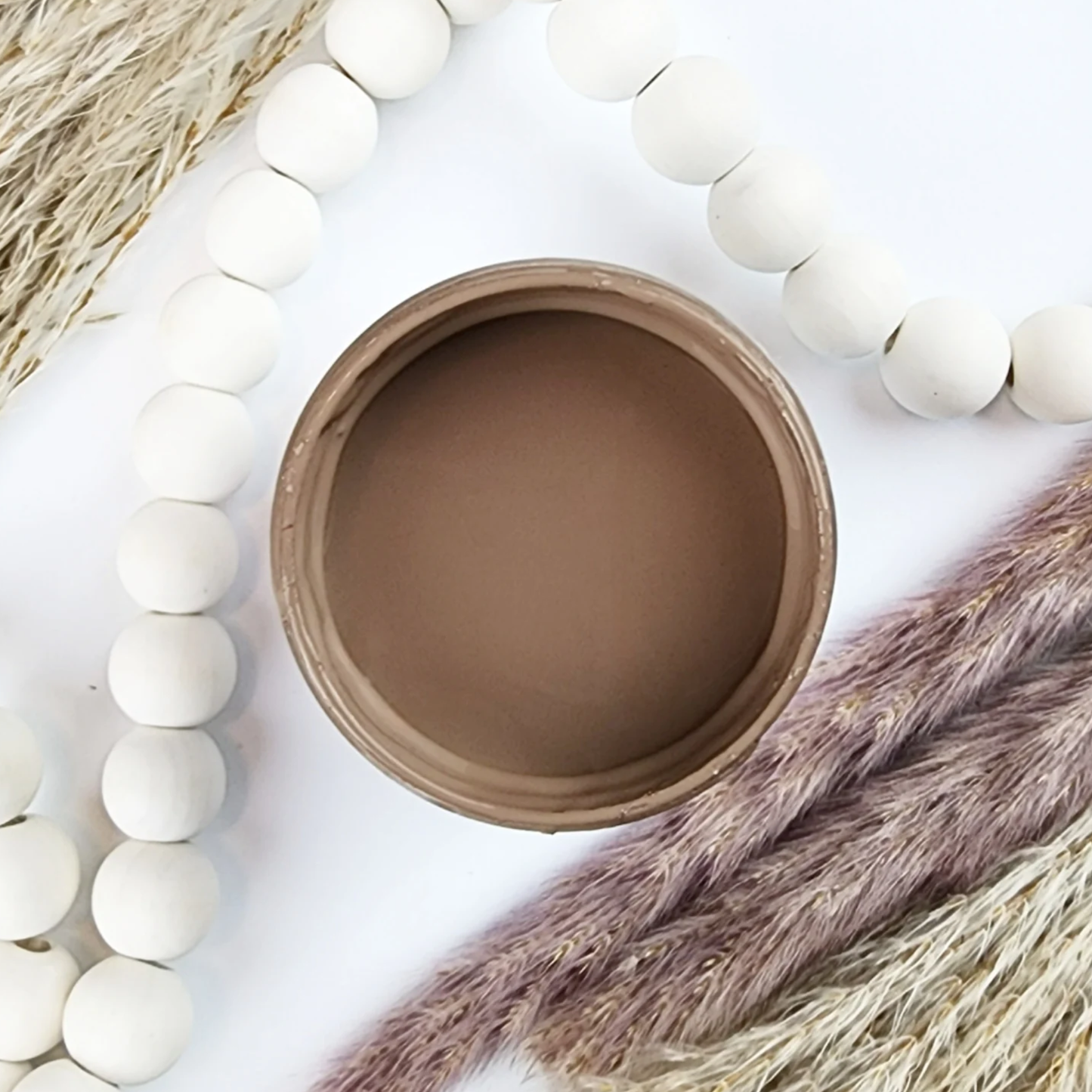 Image of open jar of cocoa colored paint with sprigs of wheat and white beads on a white background.