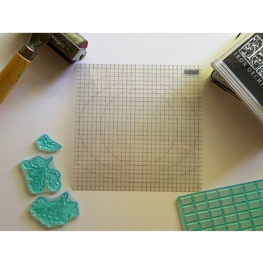 A flat-lay view of a stamping setup on a white surface, featuring a square gridded IOD Mini Thin Mount, three translucent aqua leaf-shaped stamps, a wooden-handled brayer tool, and an Iron Orchid Designs ink pad.