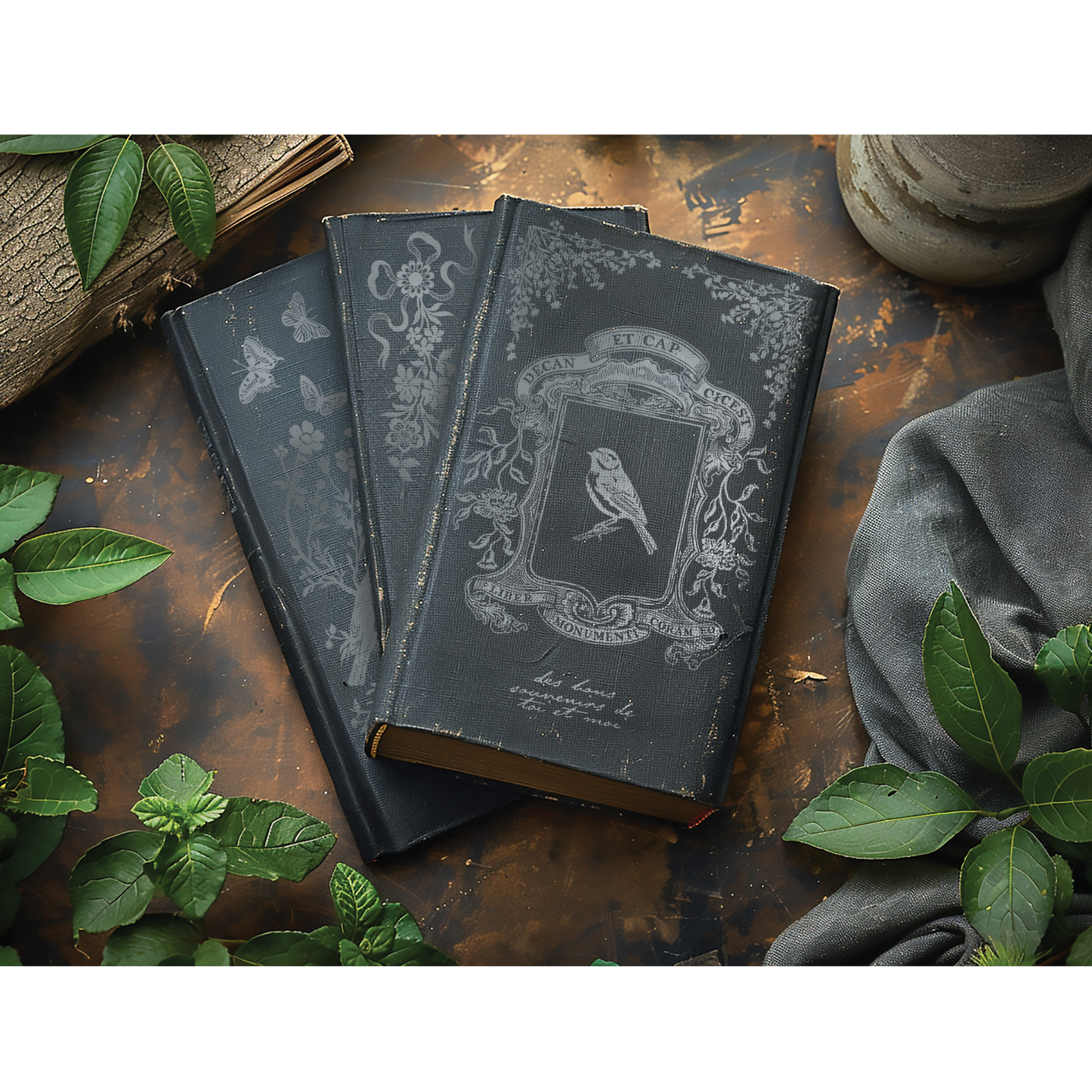 Three darkly colored books with ornate designs on a rustic wooden surface with green leaves.