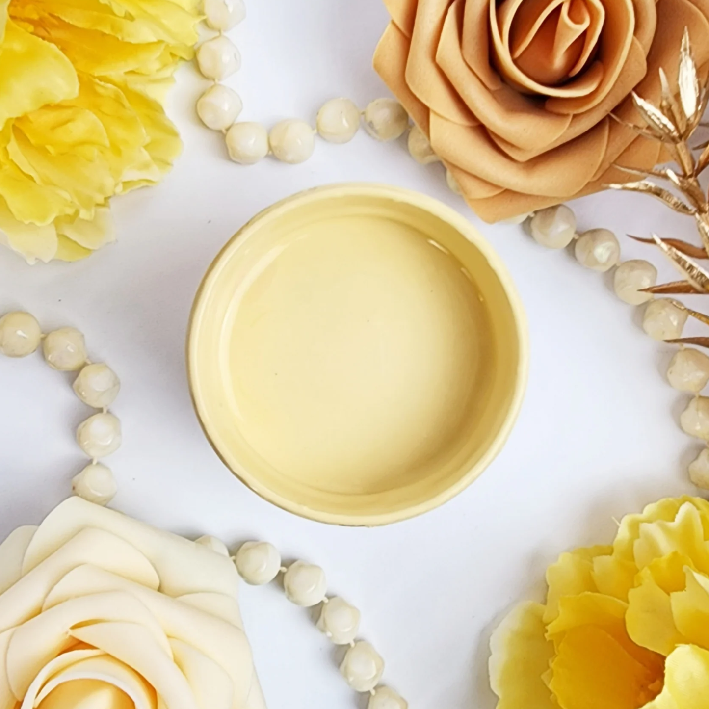 Image of open jar of light yellow-orange paint on a backround with a string of pearls and yellow and light orange flowers.