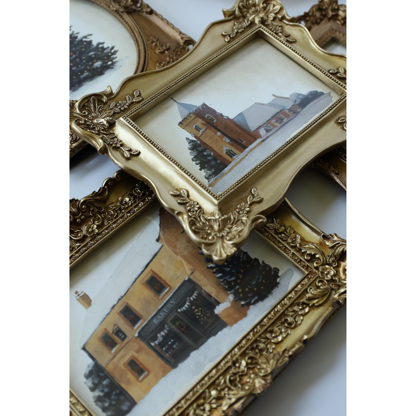 A close-up of ornate gold picture frames with carved decorative details, some containing painted architectural scenes, arranged on a light background.