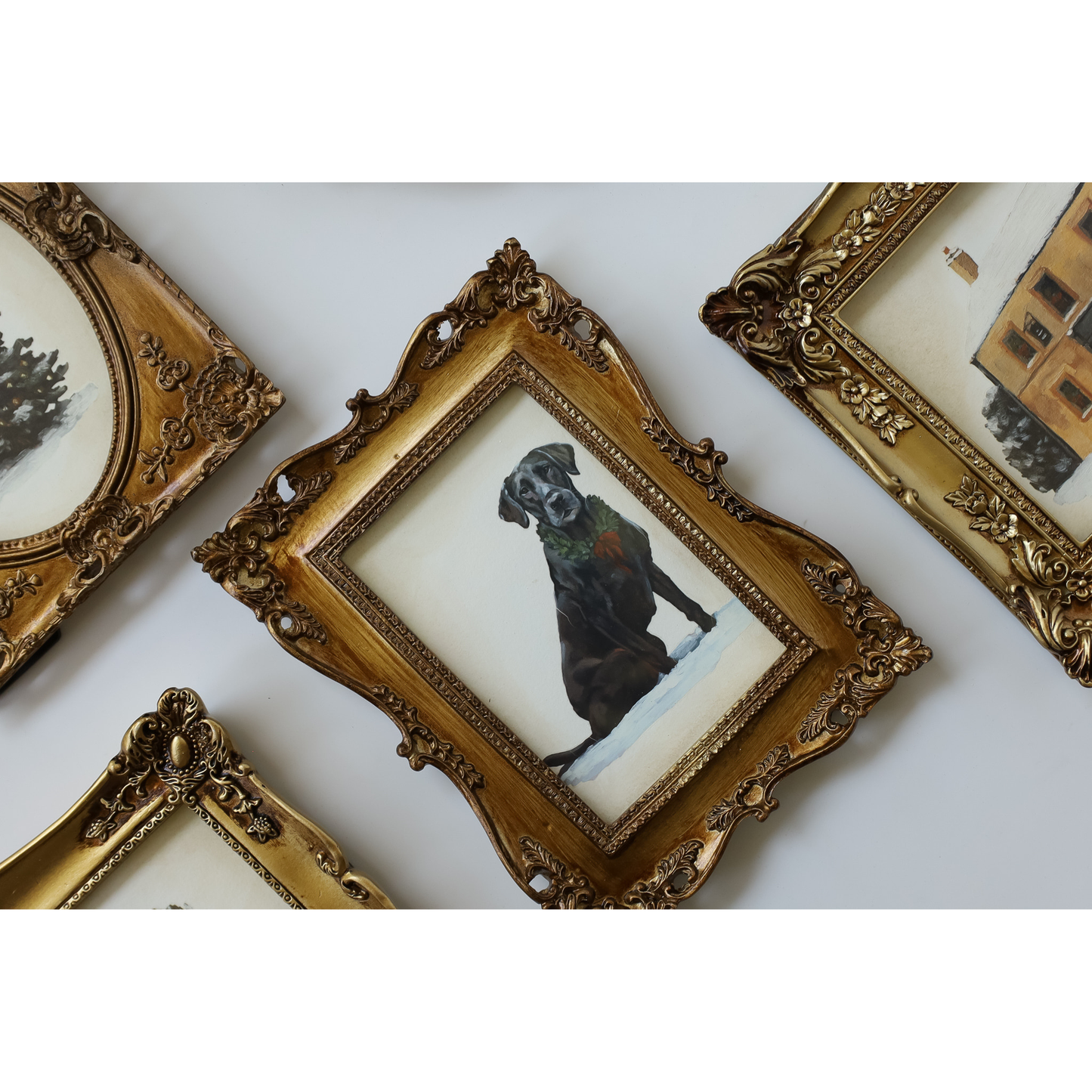 An ornate gold-framed miniature painting of a black dog in a snowy landscape, displayed on a white wall alongside other vintage frames.