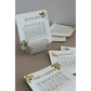 A set of decorative monthly calendar cards displayed on a gray stand, with additional cards fanned out behind and below. The cards are white with vintage-style printed grids, botanical illustrations, and gold-toned month titles including January, August, November, and September.
