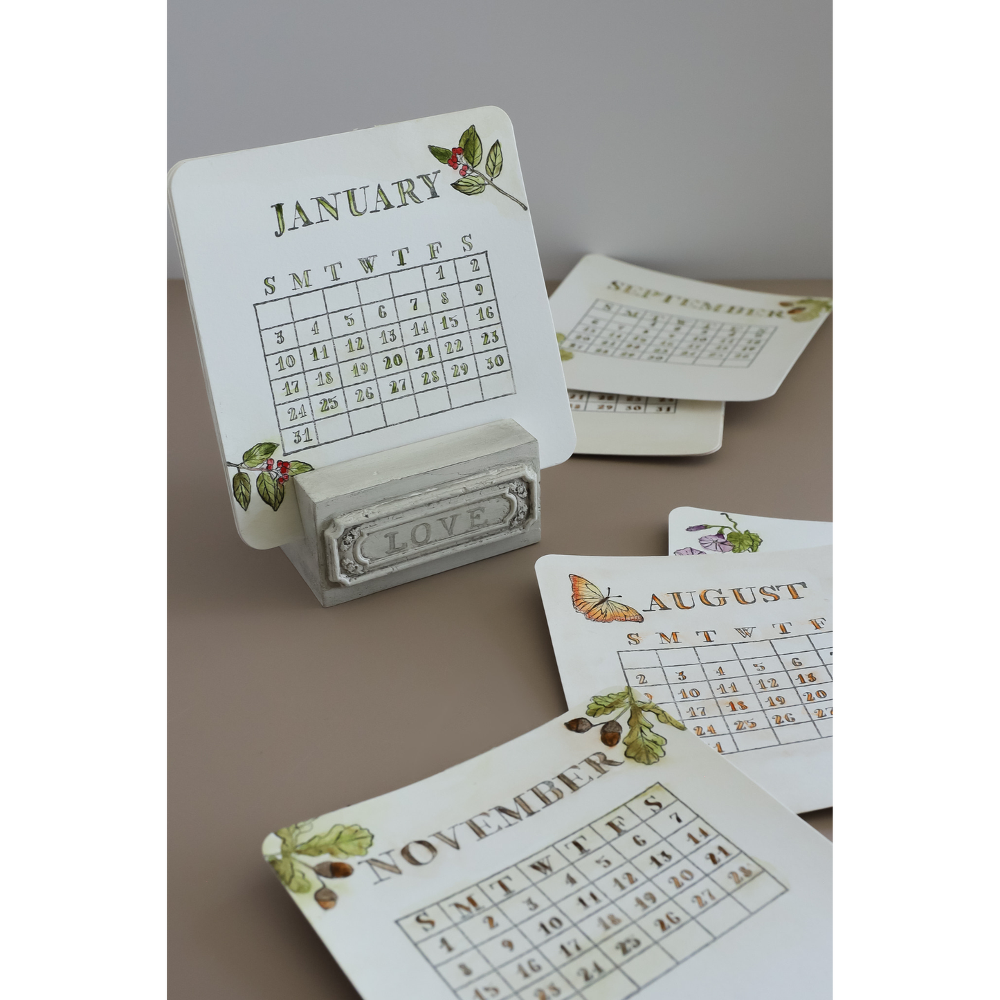 A set of decorative monthly calendar cards displayed on a gray stand, with additional cards fanned out behind and below. The cards are white with vintage-style printed grids, botanical illustrations, and gold-toned month titles including January, August, November, and September.