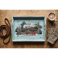 A distressed light blue wooden serving tray with a hand-painted winter cabin scene, styled on a rustic wooden tabletop with a cup of coffee, gloves, and a leather strap.