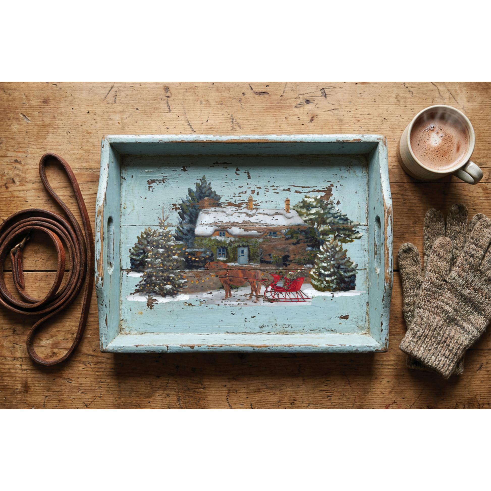 A distressed light blue wooden serving tray with a hand-painted winter cabin scene, styled on a rustic wooden tabletop with a cup of coffee, gloves, and a leather strap.