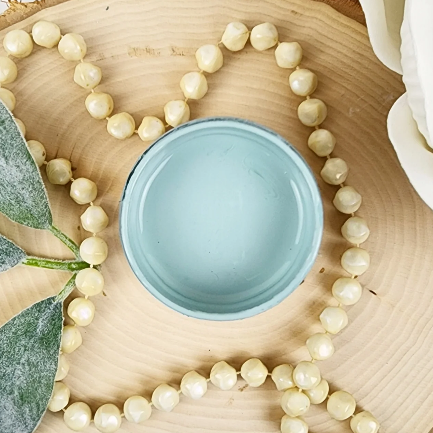 Image of open jar of pale blue paint with beige beads, leaves and white rose on a wood background.