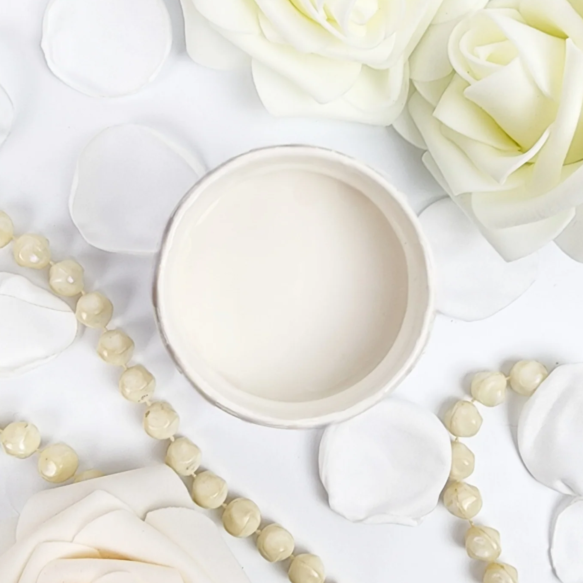 Image of open jar of creamy white paint with white roses, petals and beige beads on a white background.