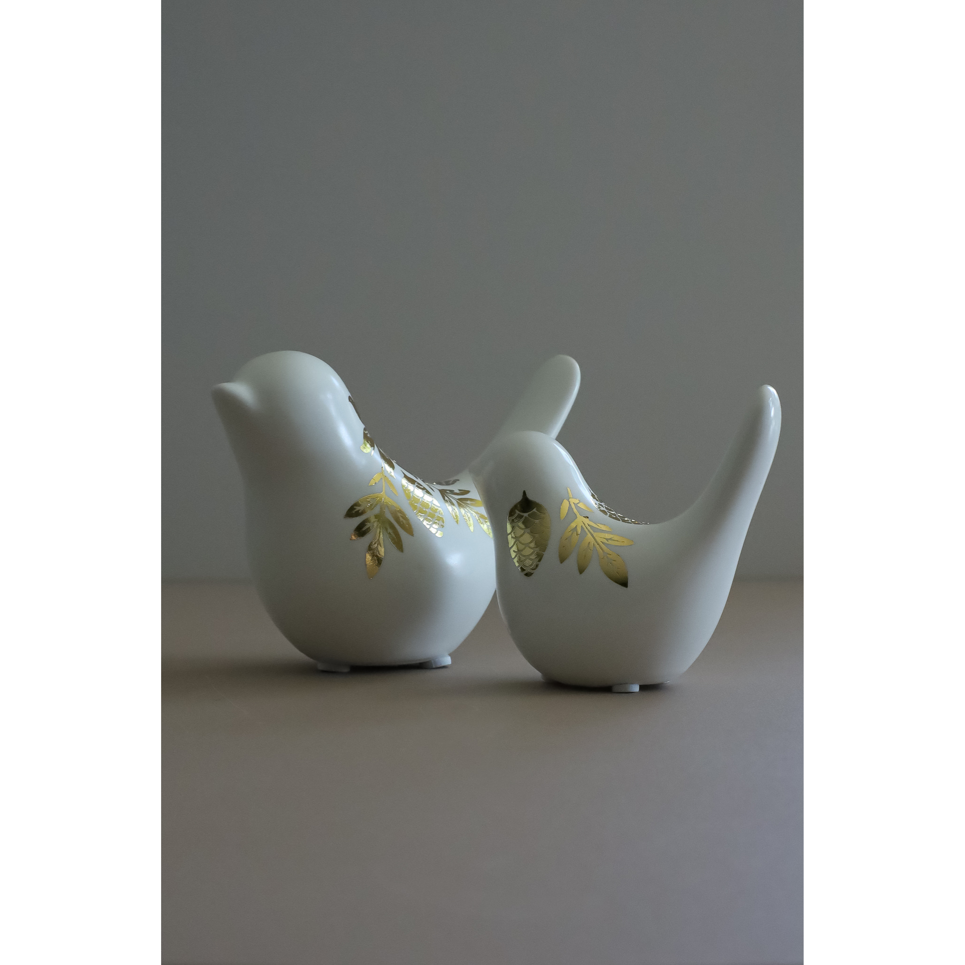 Two white ceramic bird-shaped candle holders with gold image transfer accents, standing side by side on a neutral surface against a gray background.