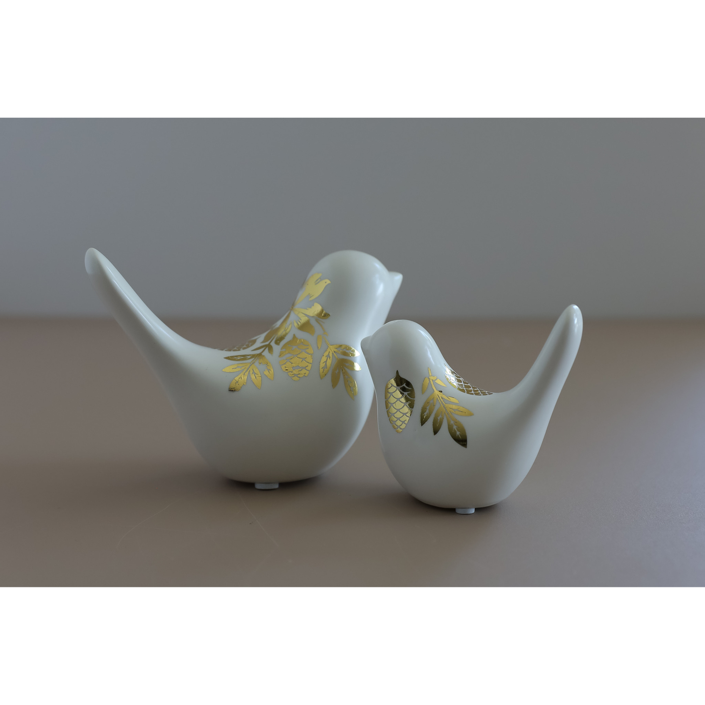 Two white ceramic bird figurines with gold image transfer pinecone designs, displayed on a neutral beige surface against a gray background.