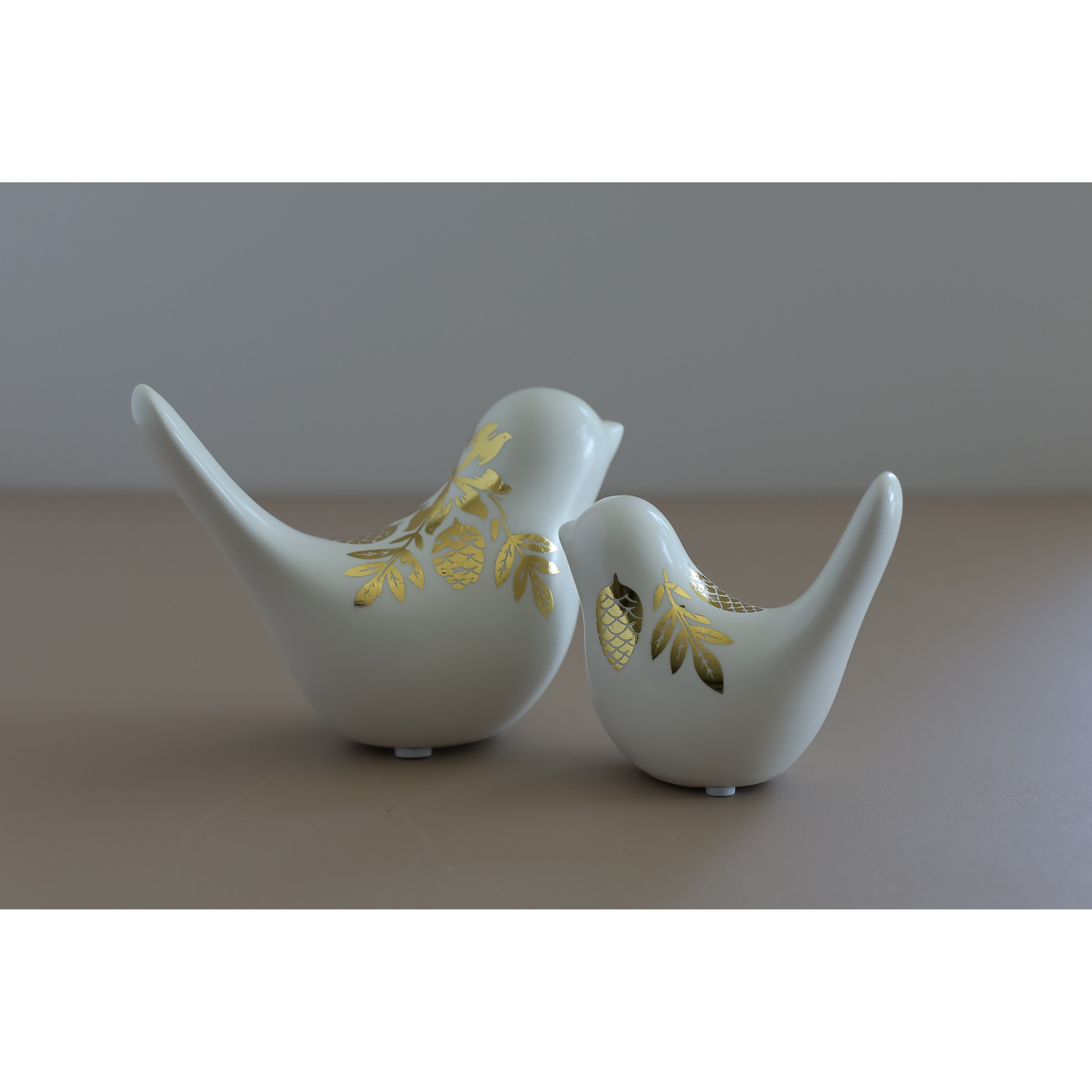 Two white ceramic bird figurines with gold image transfer pinecone designs, displayed on a neutral beige surface against a gray background.