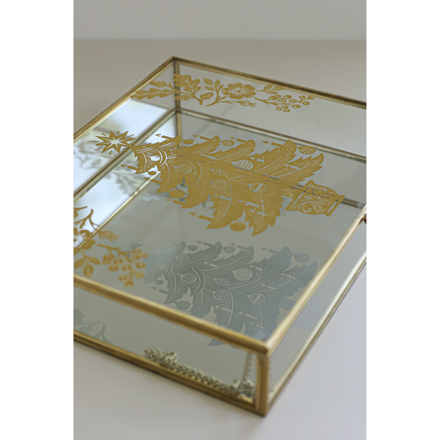A rectangular glass jewelry box with a polished gold metal frame and gold floral transfers on the glass, shown on a light neutral background.