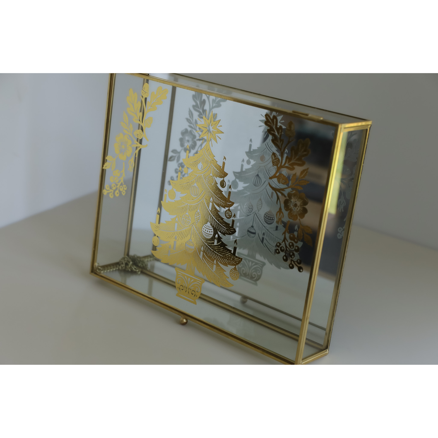 A rectangular glass jewelry box with a gold metal frame, featuring gold and silver Christmas tree and floral transfer designs on the mirrored lid, set against a neutral light gray background.