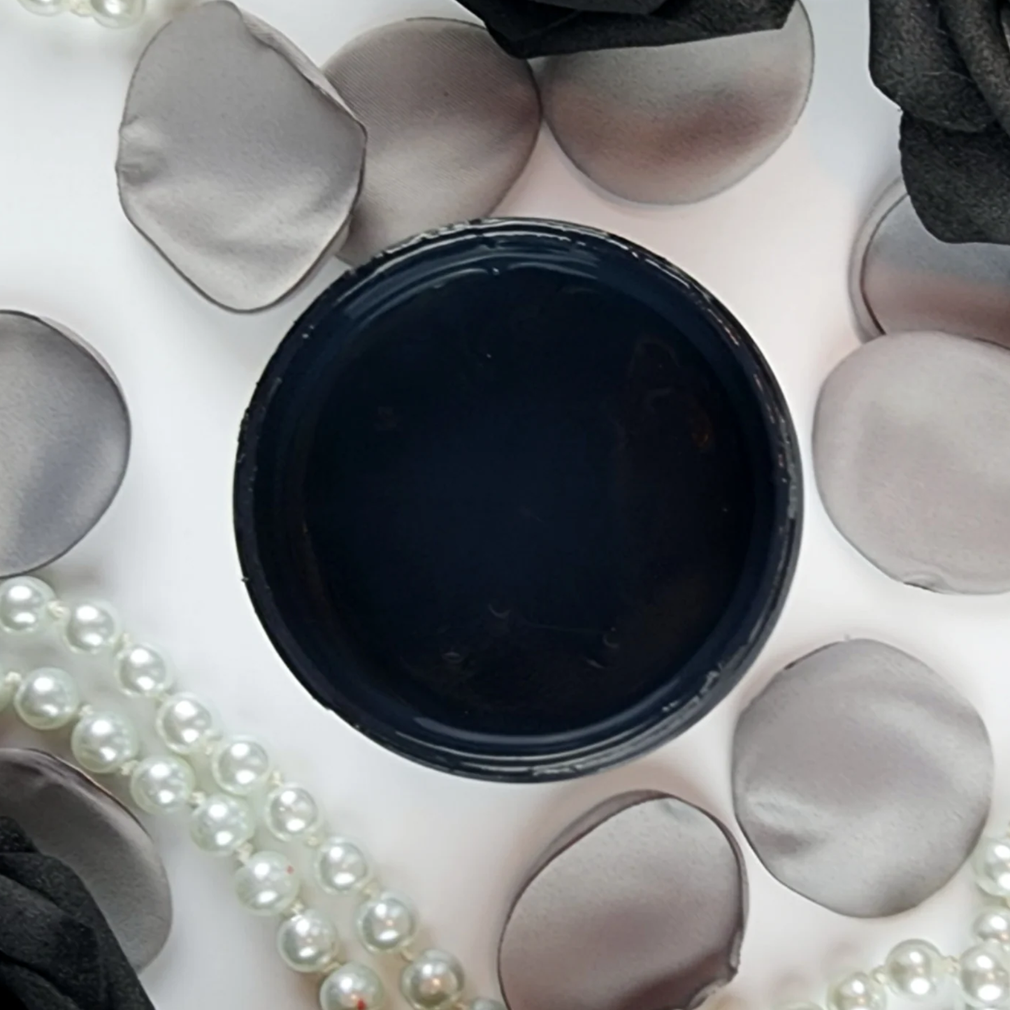 Image of open jar of black paint with oyster and beige colored silk petals and pearls on a white background.