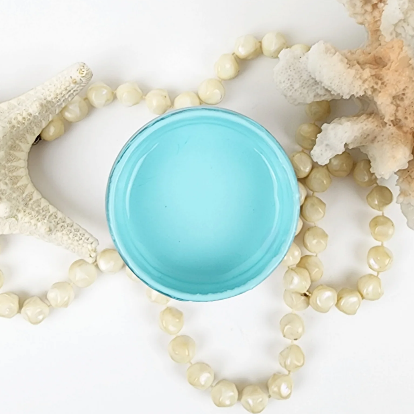 Image of open jar of light turquoise paint with coral, starfish and beige beads on a white background.