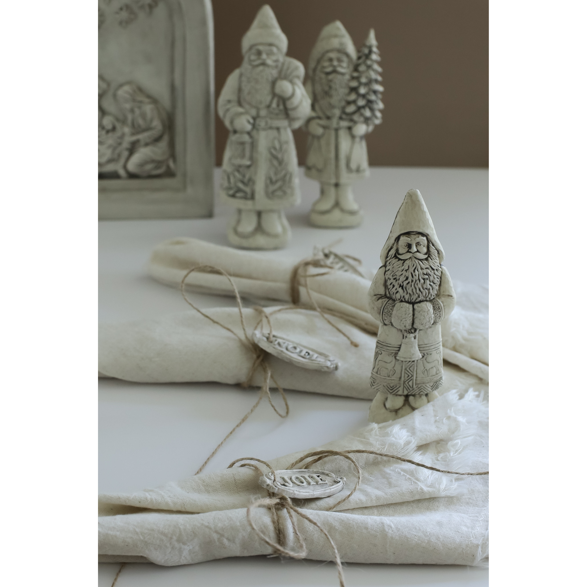 A set of rustic, hand-painted Santa Claus figurines in a light gray stone-like finish, styled with cream-colored linen napkins tied with twine and small silver-toned metal tags on a neutral tabletop.
