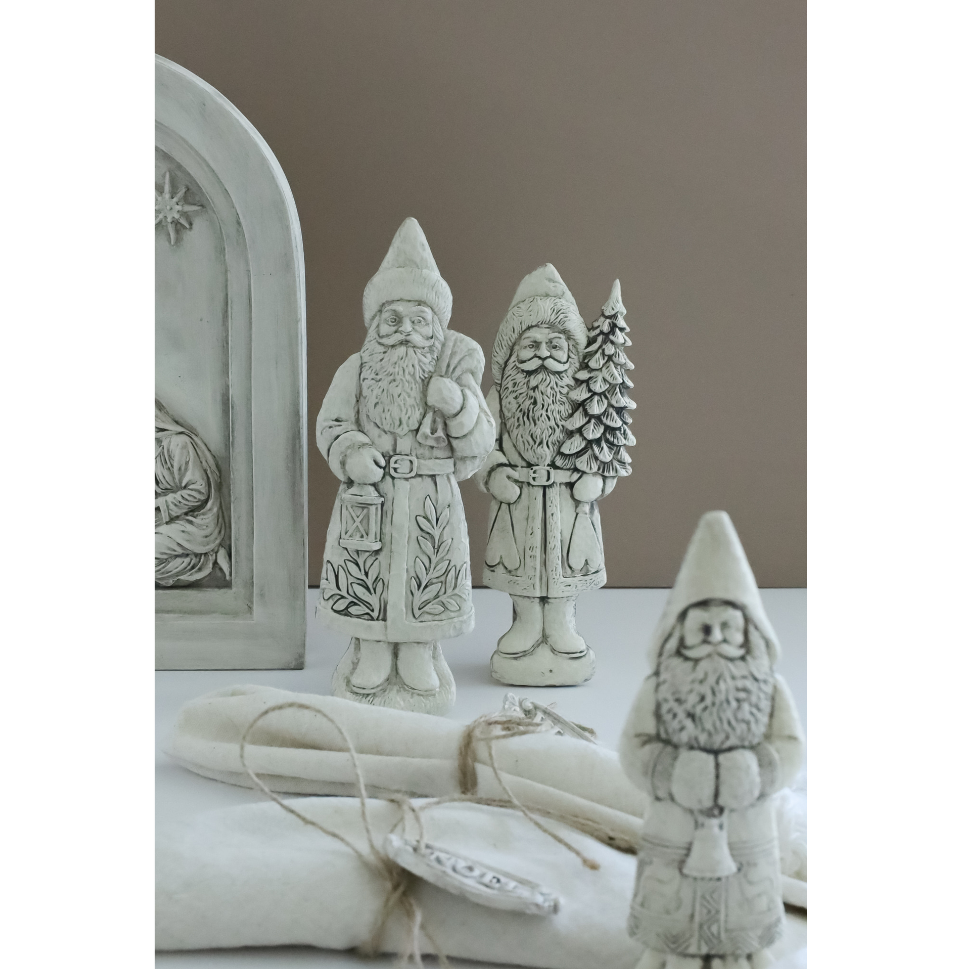 A set of rustic white clay Santa Claus figurines, including a large arched window ornament, a standing Santa, a Santa holding a Christmas tree, and a smaller Santa in the foreground, arranged on a white surface with a neutral brown background.