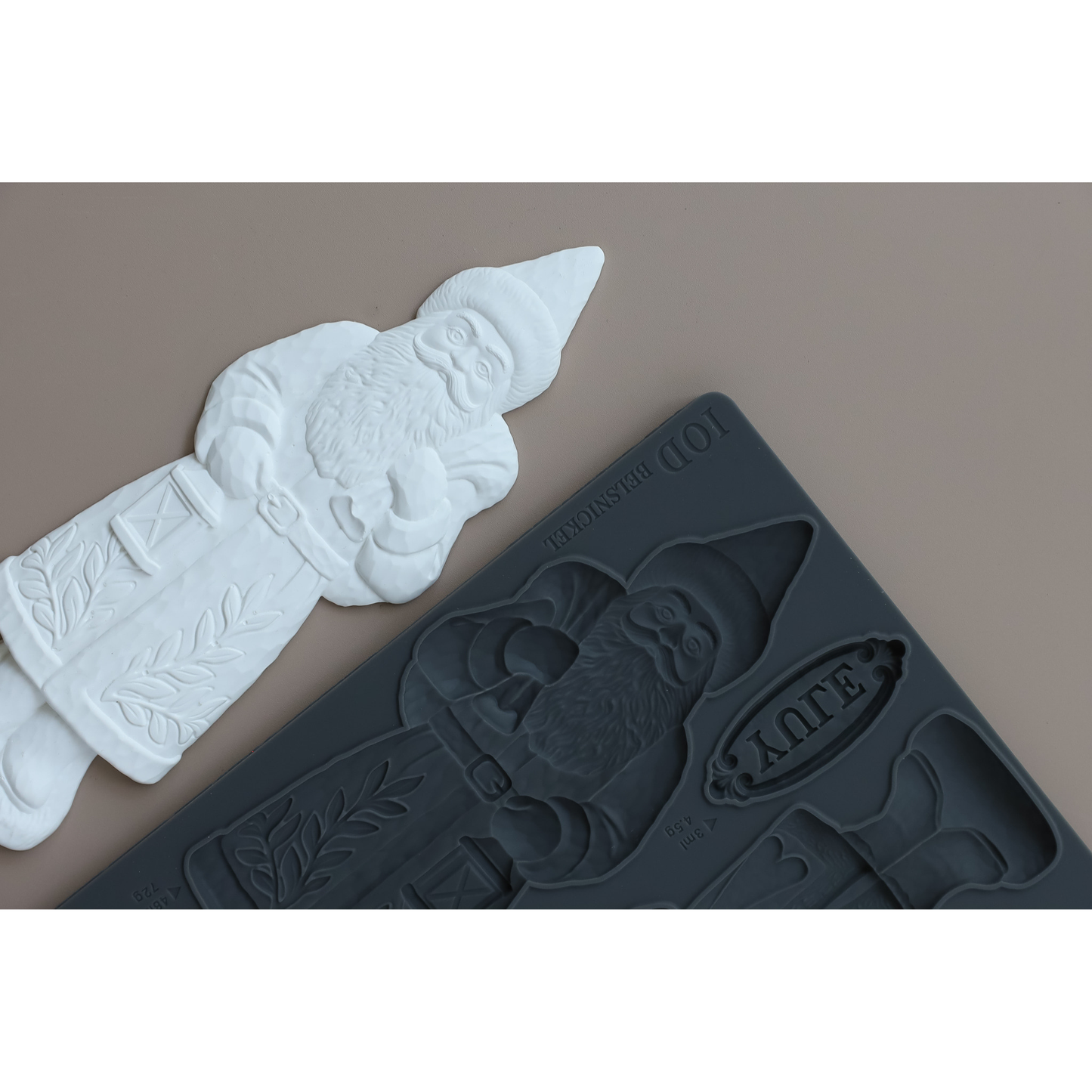 A dark gray silicone mold featuring a vintage santa design, shown next to the cast figure on a solid taupe background.