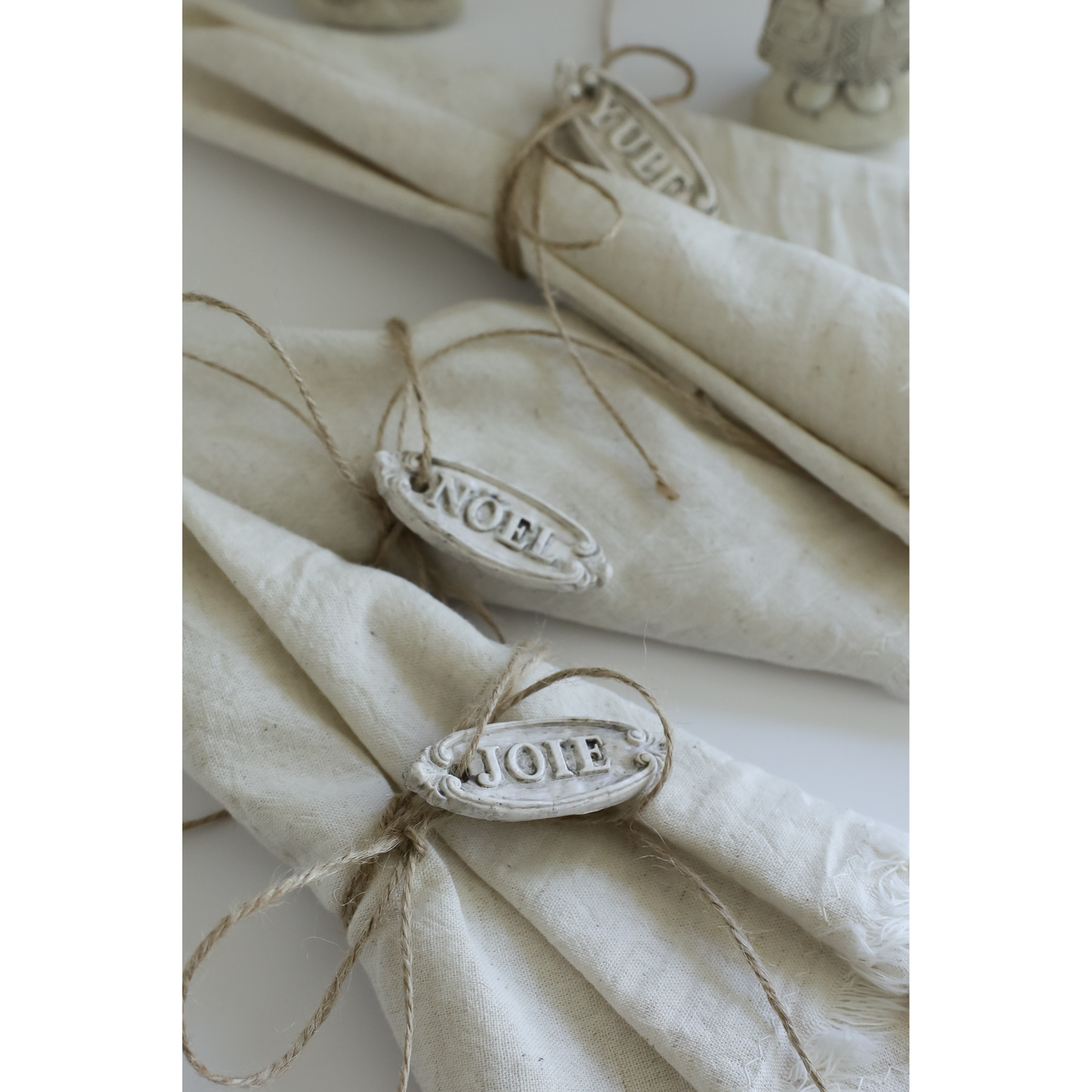 Three cream-colored linen napkins tied with twine and decorated with oval white metal place cards engraved with 'JOIE' are arranged on a white surface.