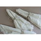 Three folded white linen napkins with frayed edges, tied with twine and decorated with silver metal tags engraved with 'JOIE' and 'YULE' arranged on a solid taupe background.