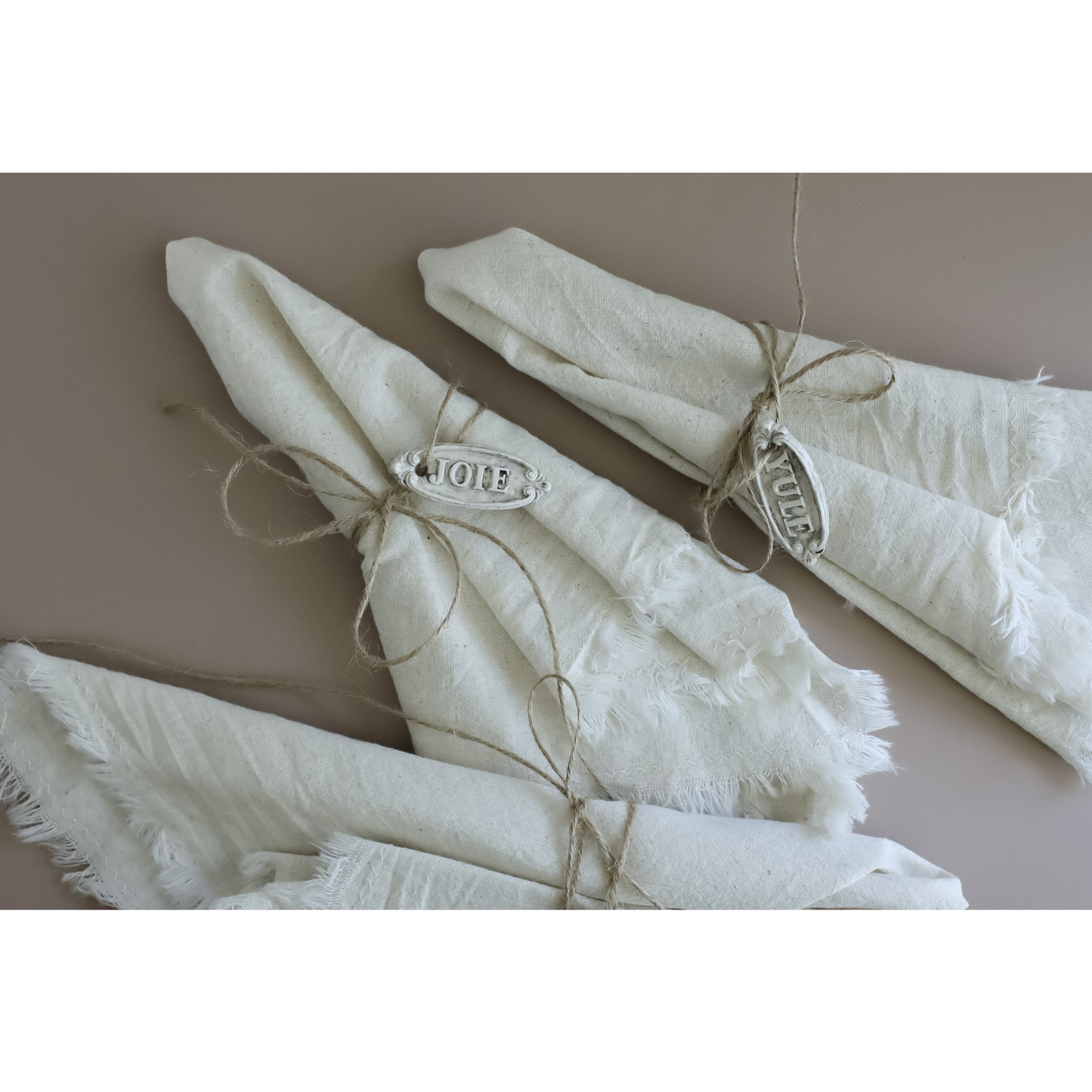 Three folded white linen napkins with frayed edges, tied with twine and decorated with silver metal tags engraved with 'JOIE' and 'YULE' arranged on a solid taupe background.
