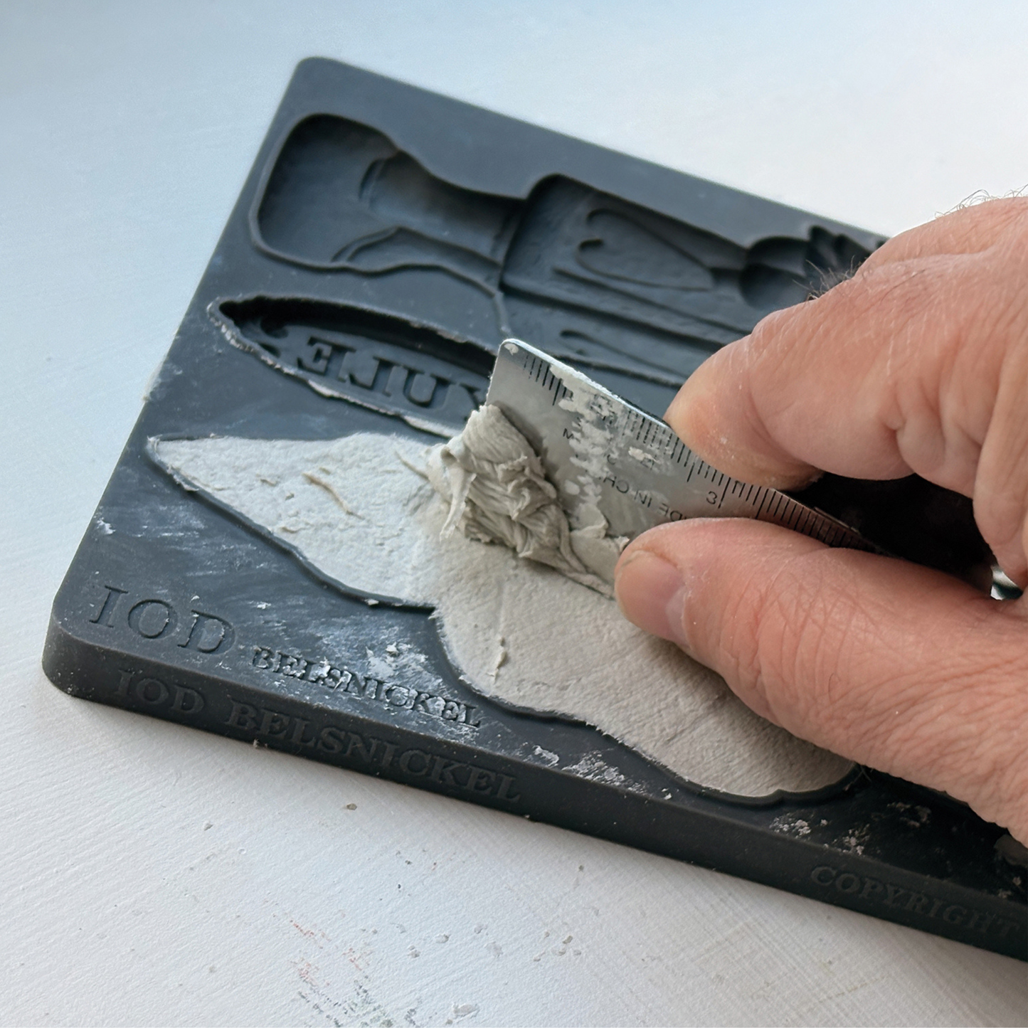 A close-up of a hand using a small metal ruler to spread clay into a silicone mold, with embossed text including 'IOD BElSNICKEL' visible on the mold.