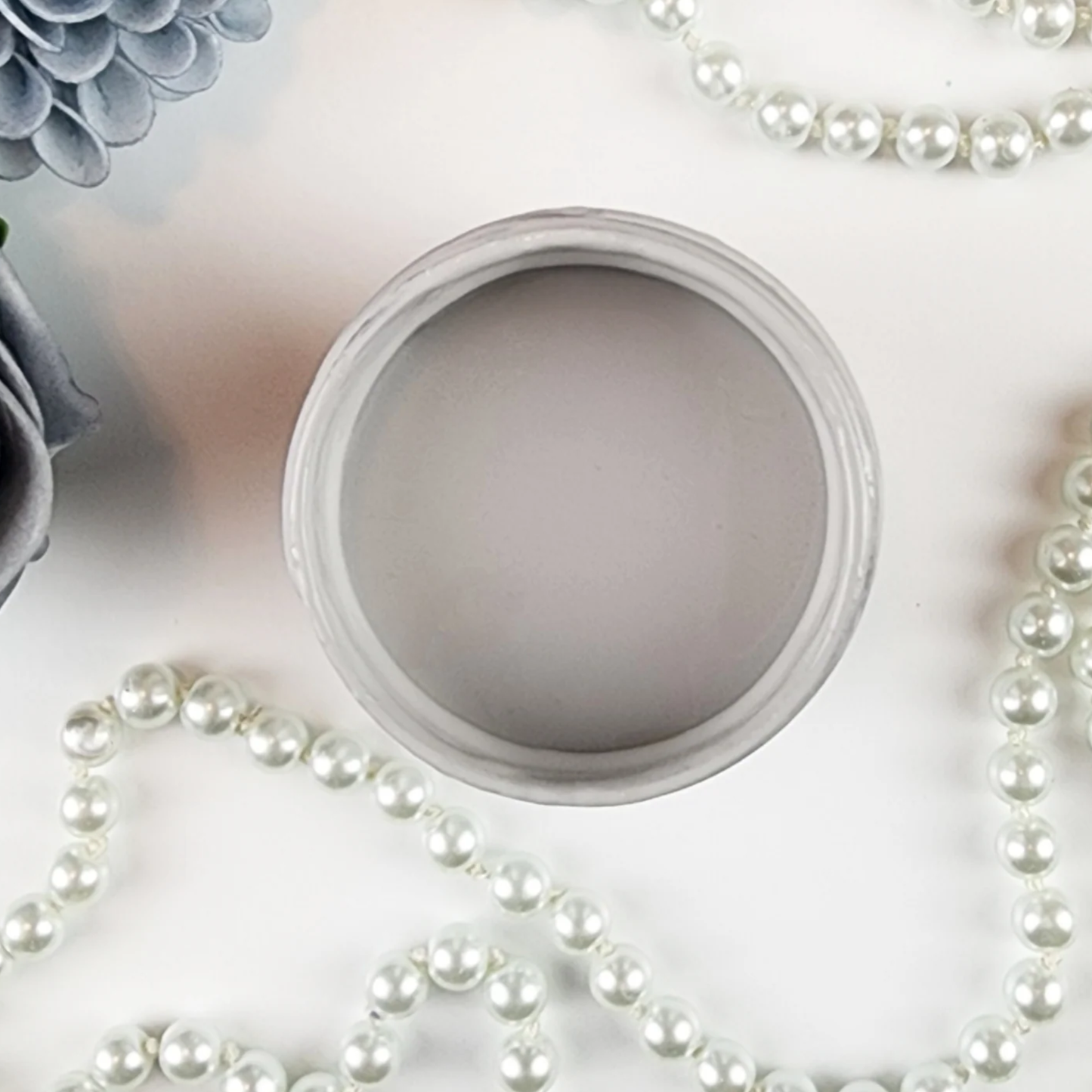 Image of open jar of light taupe/grey colored paint with pearls and blue flower on a white background.