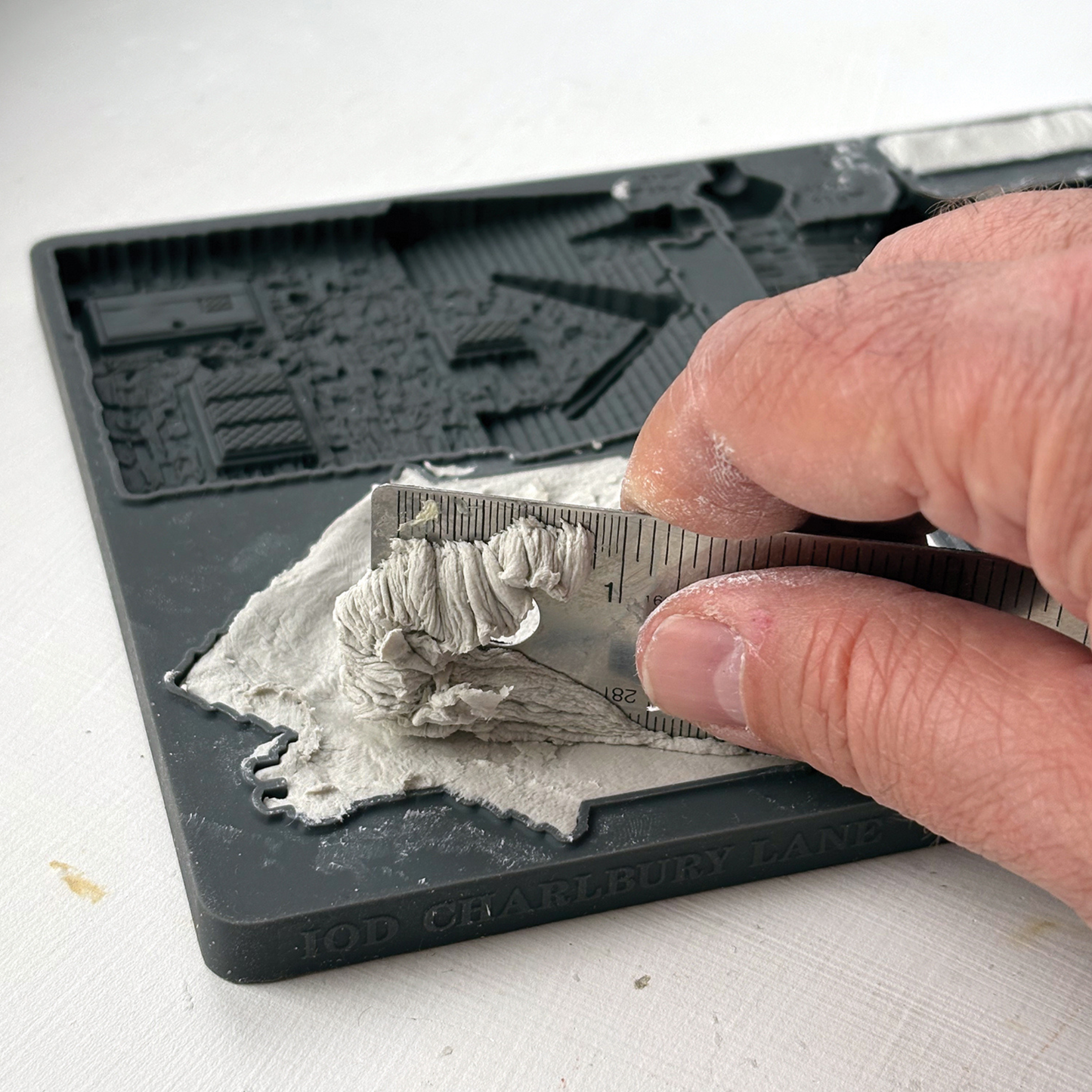 A close-up of a hand using a metal ruler to spread level clay on a silicone mould.
