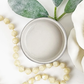 Image of open jar of beige/taupe colored paint with leaves, white rose and beige beads on a white background.