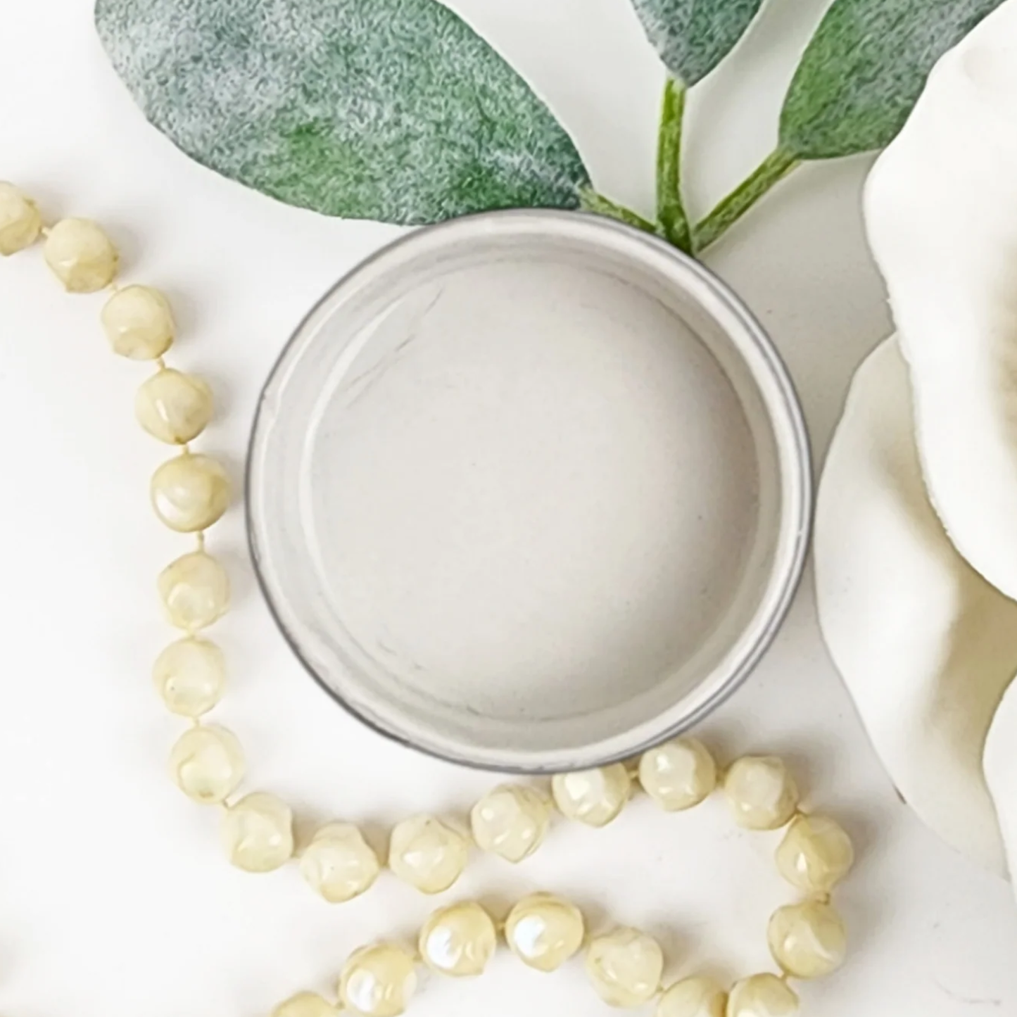 Image of open jar of beige/taupe colored paint with leaves, white rose and beige beads on a white background.