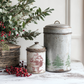 "Christmas Wishes" IOD Stamp by Iron Orchid Designs. Single sheet stamp. Available at Milton's Daughter. Example pic. Two vintage-style metal canisters with decorative lids on a wooden surface with greenery and berries.