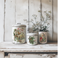 "Christmas Joy" IOD Transfer by Iron Orchid Designs. Available at Milton's Daughter. Pic 2. Three vintage-style ceramic canisters with decorative lids on a wooden surface.