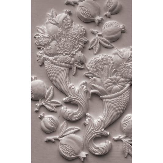 A close-up of a white castng featuring a classic fruit and floral cornucopia motif, including pomegranates, cherries, and bouquets, set against a matching textured background.