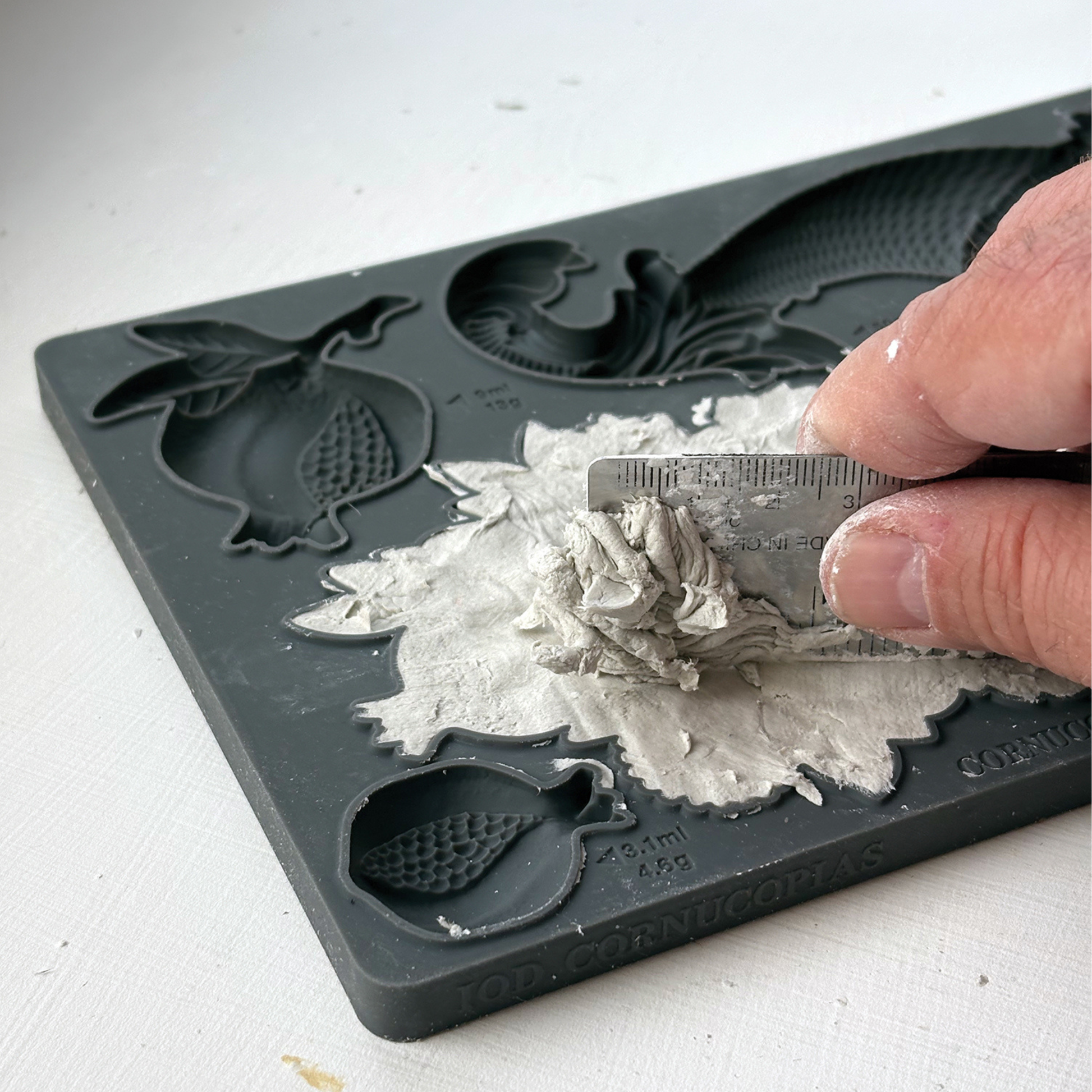 A close-up of a hand using a small metal ruler to press and shape wet clay or plaster into a decorative relief on a dark gray silicone mold tray.