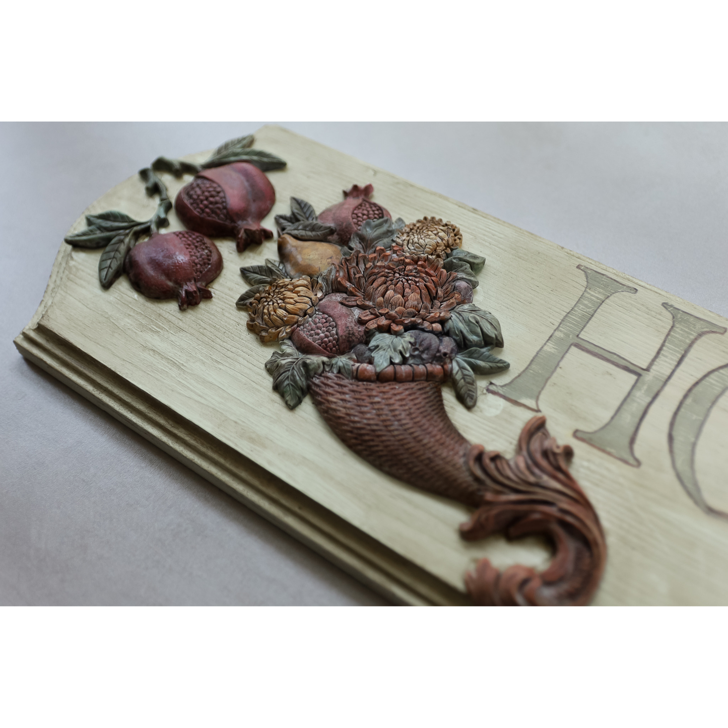 A close-up of a decorative carved wooden plaque featuring pomegranates, flowers, and a basket motif, with part of a large letter 'H' visible on the right, set against a neutral light gray background.