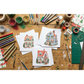 Three printed cards featuringIOD transfers are arranged on a wooden craft table surrounded by art supplies, glitter, paintbrushes, tape, and markers.
