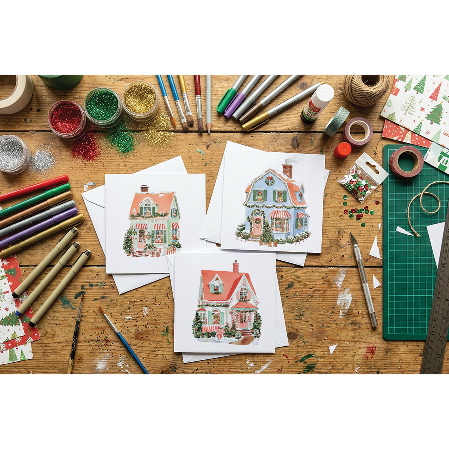 Three printed cards featuringIOD transfers are arranged on a wooden craft table surrounded by art supplies, glitter, paintbrushes, tape, and markers.