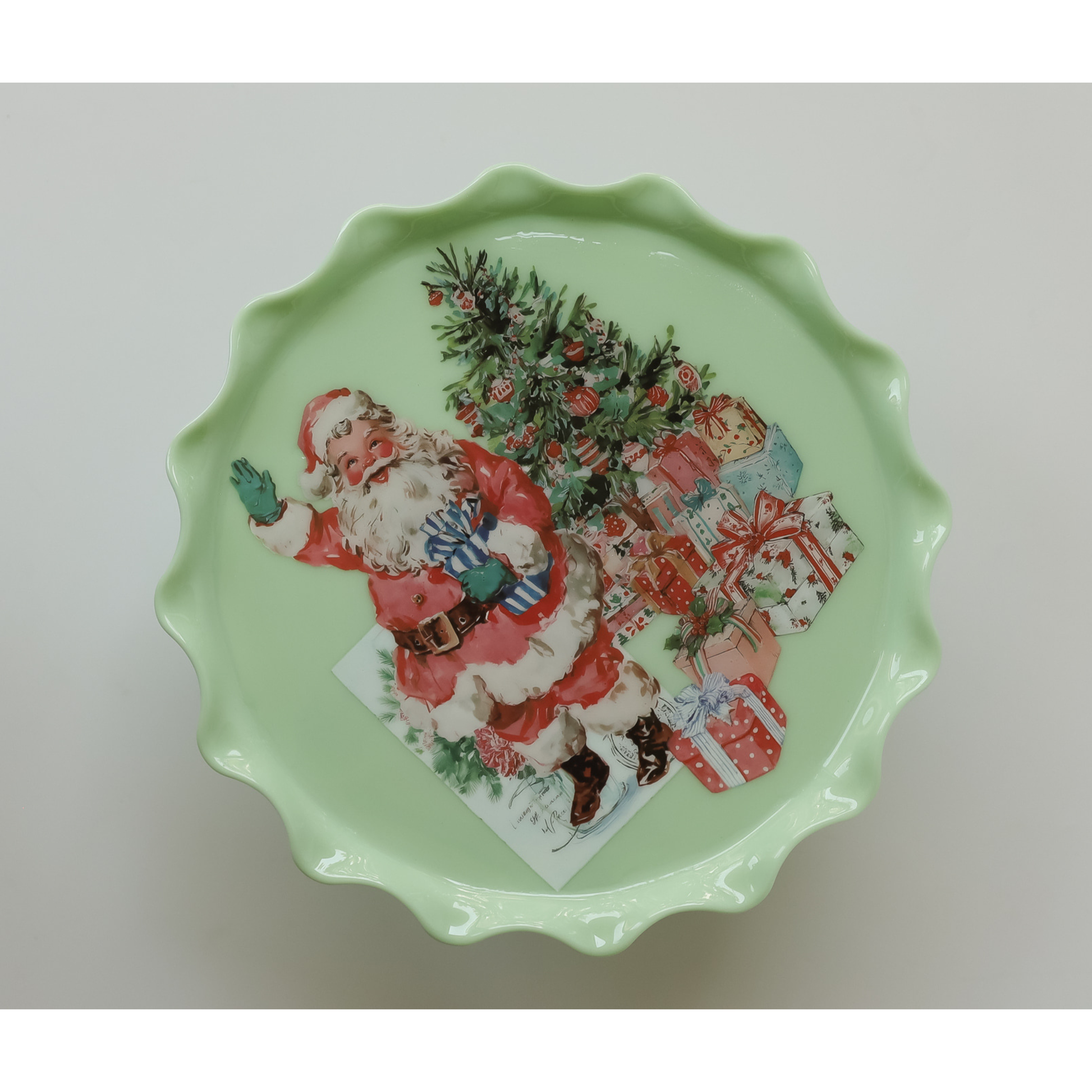 A scalloped-edge ceramic plate or dish in pale mint green, featuring a vintage-style IOD transfer image of Santa Claus sitting next to a decorated Christmas tree and wrapped gifts.