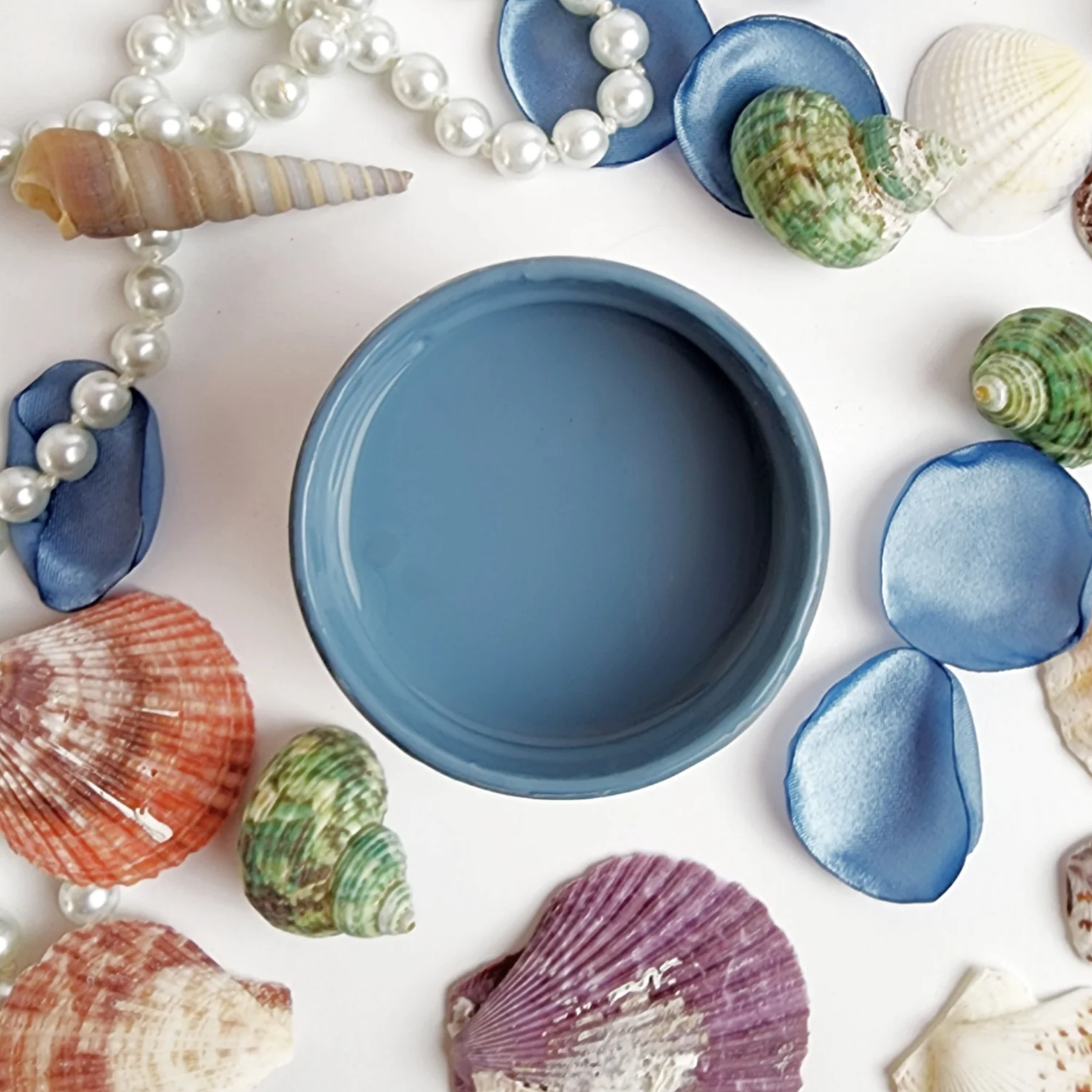 Image of open jar of medium blue paint with colorful sea shells and blue flower petals on a white background.