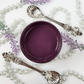 Image of open jar of deep purple colored paint with pearls and silver spoons on a white background.