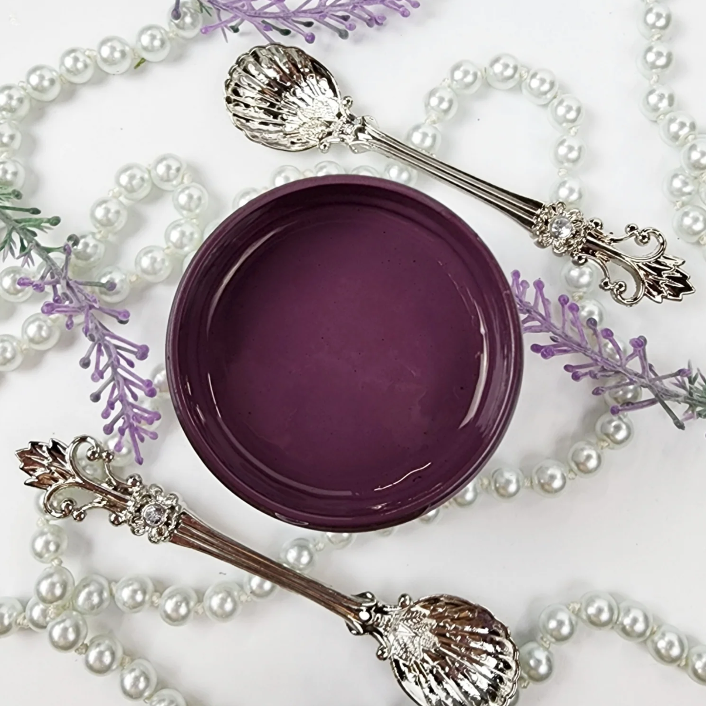 Image of open jar of deep purple colored paint with pearls and silver spoons on a white background.