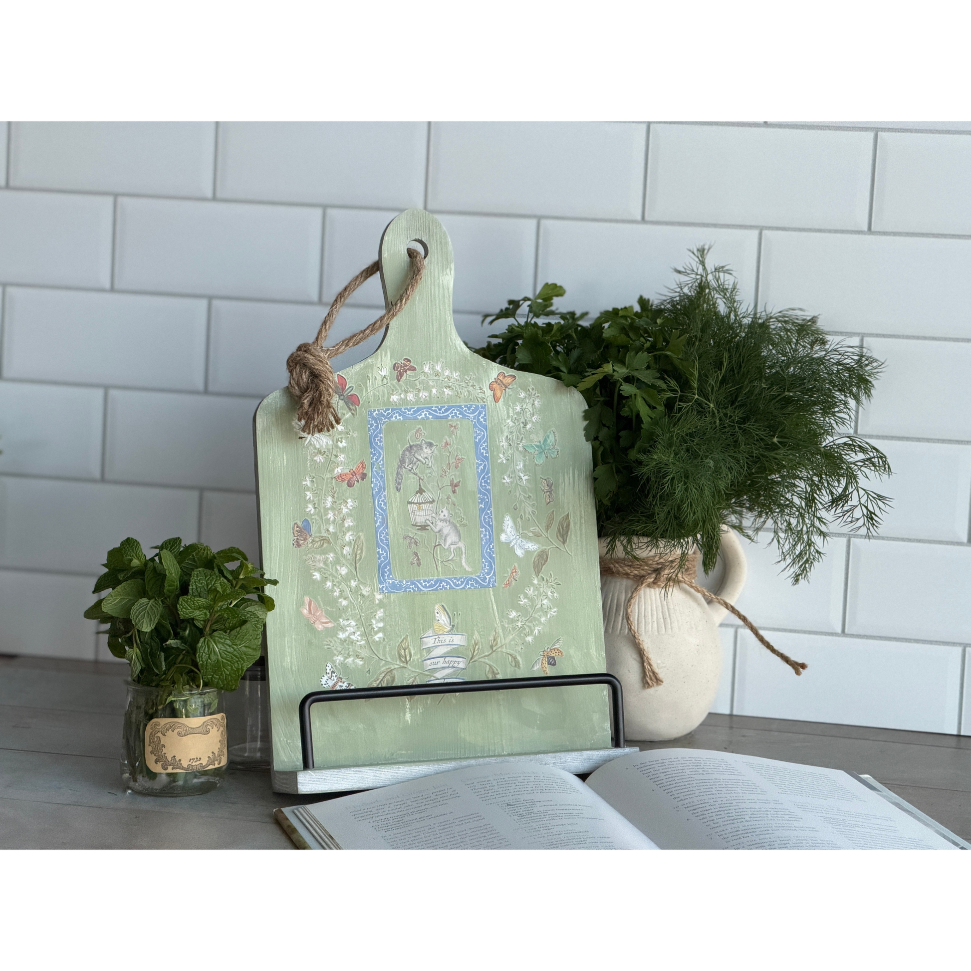 Decorative cutting board with floral patterns on a kitchen counter with plants and an open book.