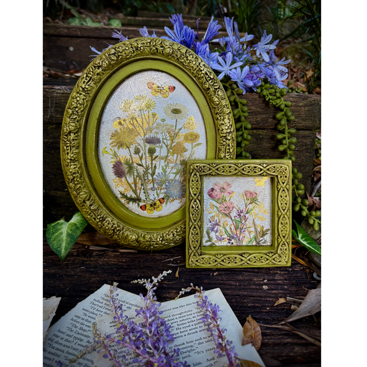 Two decorative framed artworks with floral designs on a rustic wooden surface with flowers and an open book.