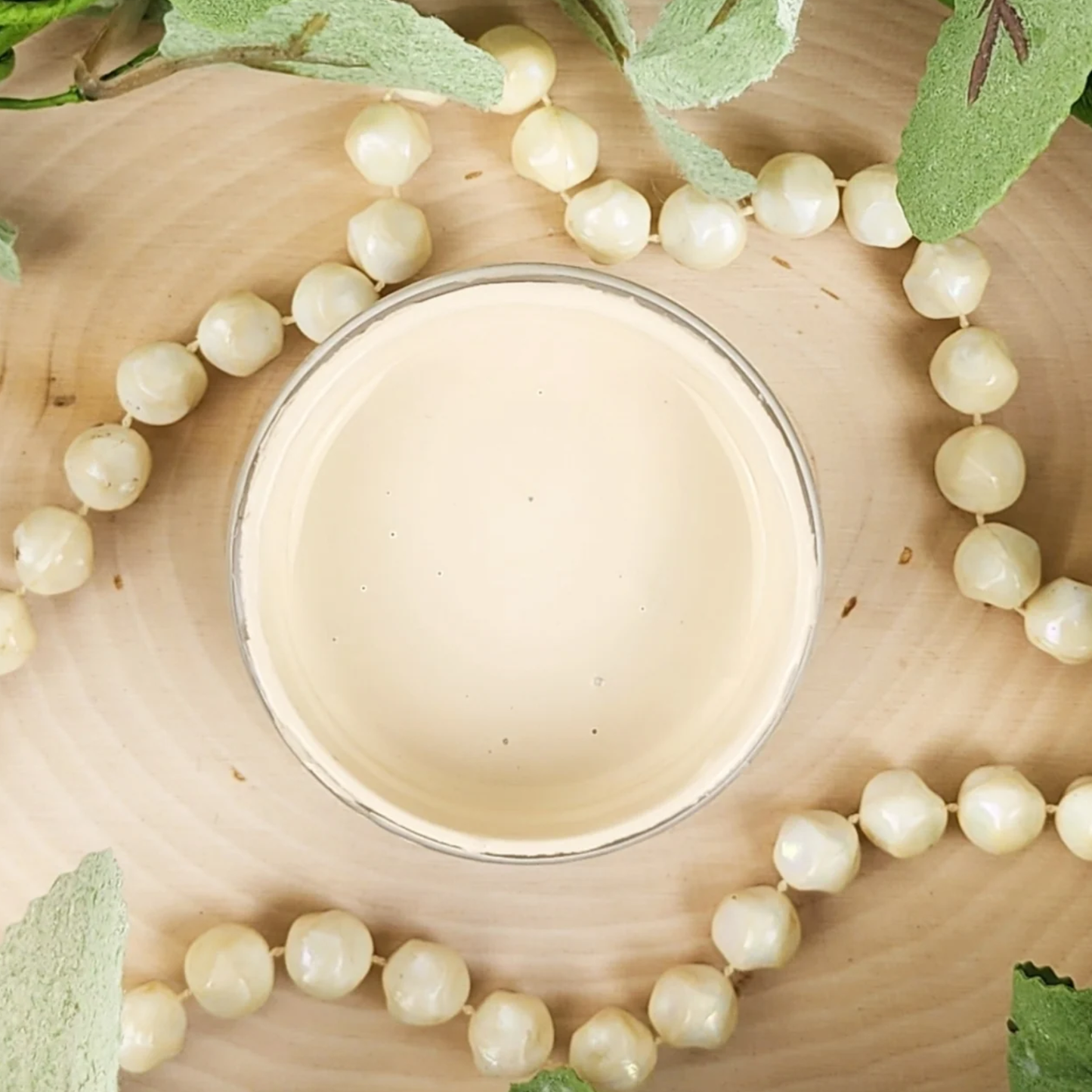 Image of open jar of light beige paint with beads and leaves on a wood background.