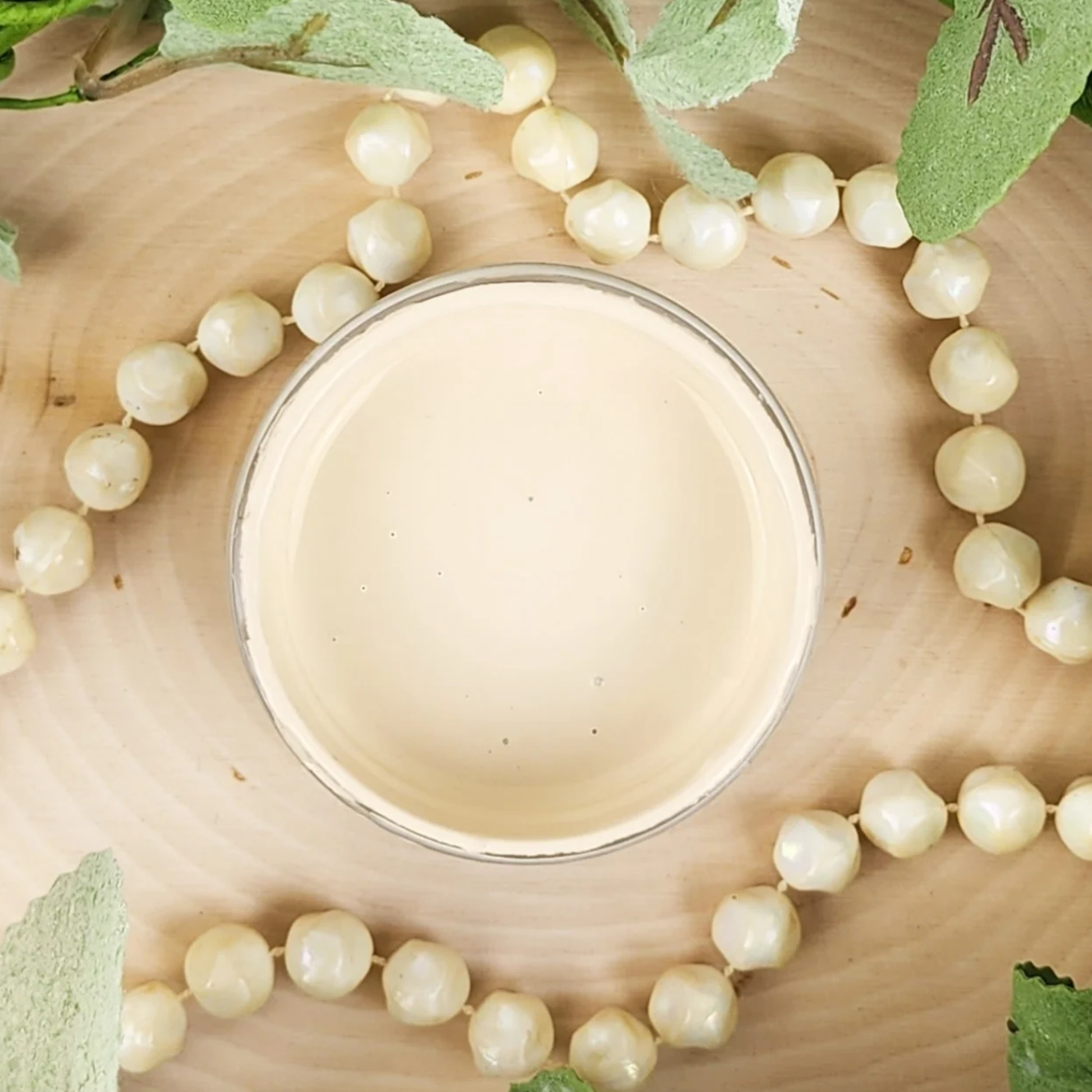 Image of open jar of light beige paint with beads and leaves on a wood background.