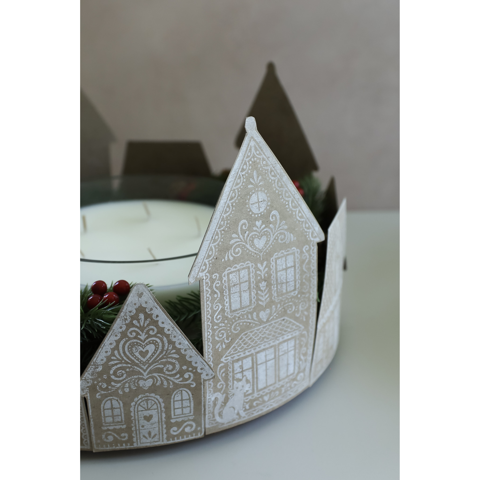 A  decorative house-shaped candle holder with intricate gingerbread-style patterns on kraft paper and wihte ink, holding a white candle and green pine garland with red berries.