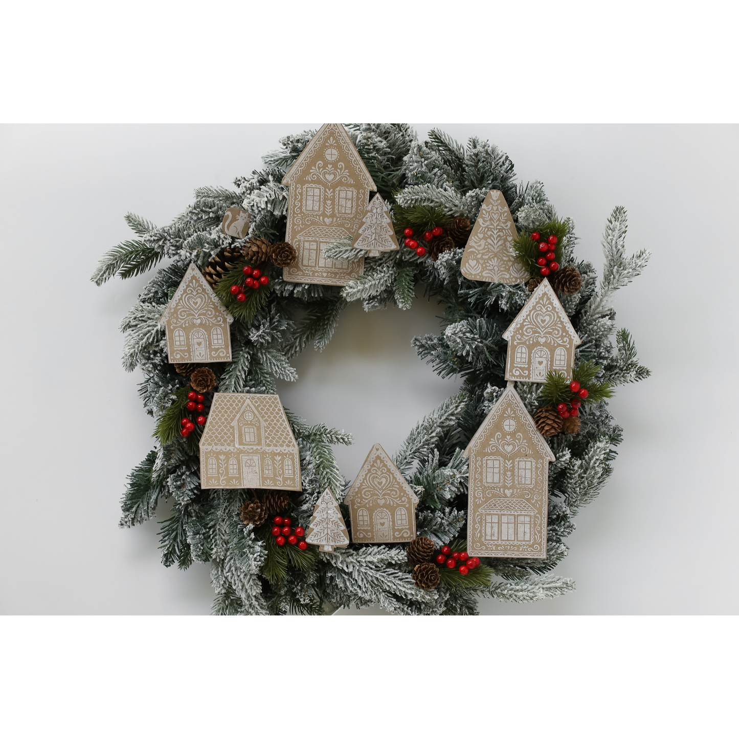 A festive Christmas wreath decorated with frosted green pine branches, small red berry clusters, pinecones, and multiple beige wooden house ornaments in different sizes, shown against a plain light gray background.